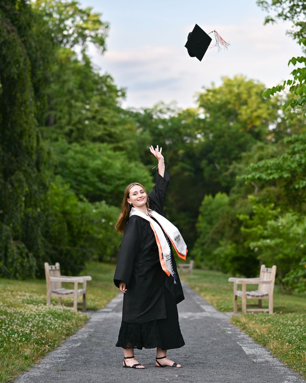 A smiling graduate in black gown tosses cap in air on tree-lined path, wearing colorful honor stole.