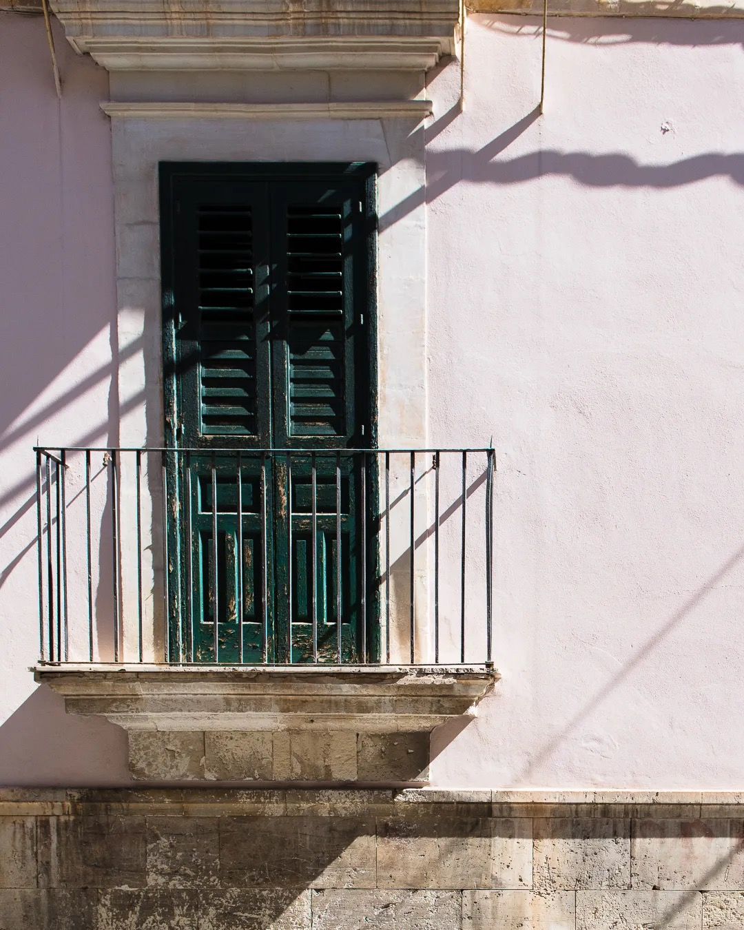 A pastel pink building features an old green shuttered balcony casting shadows in the afternoon sun.