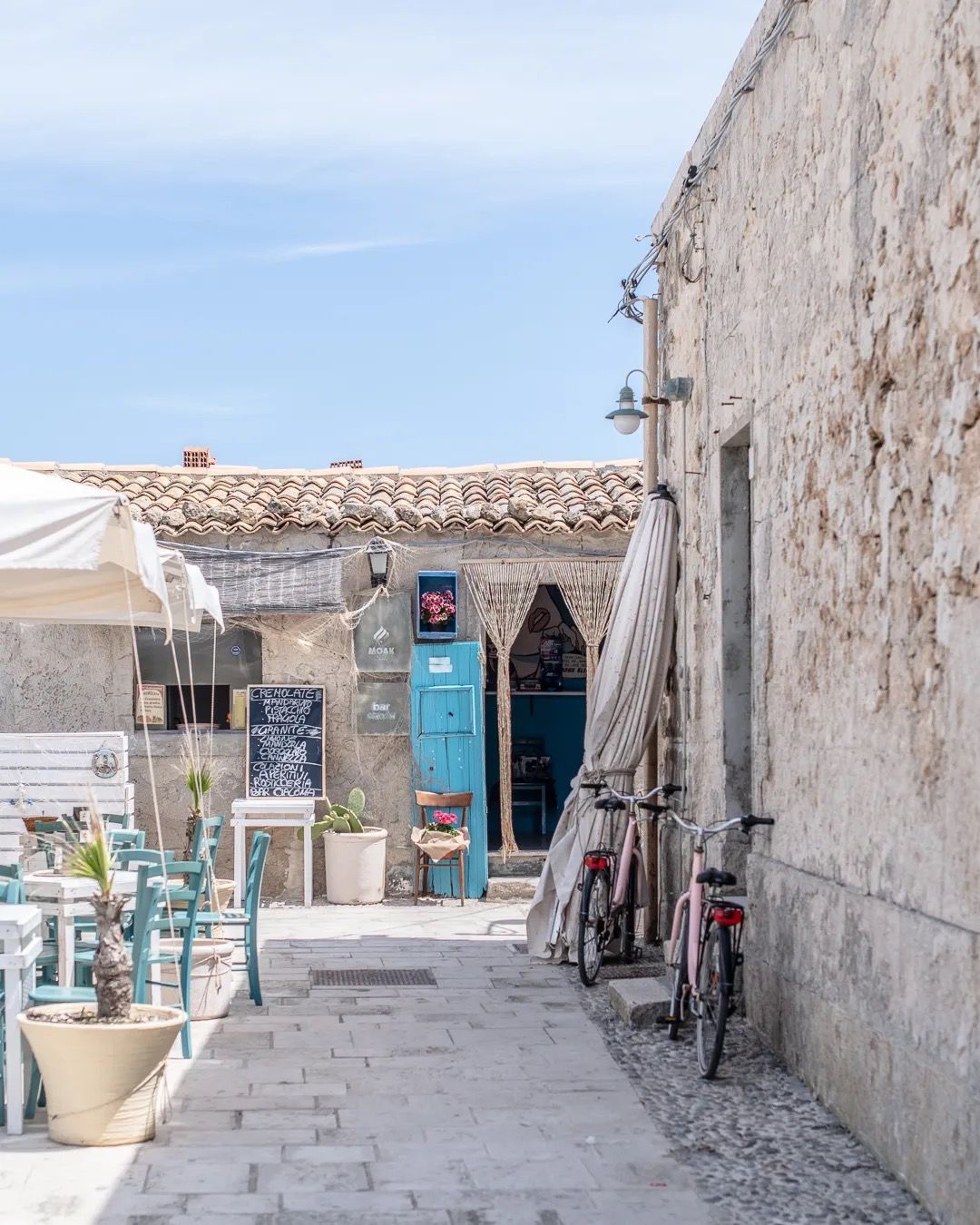 A narrow stone alley with shops and signs opens toward a bright blue sea on a sunny day.