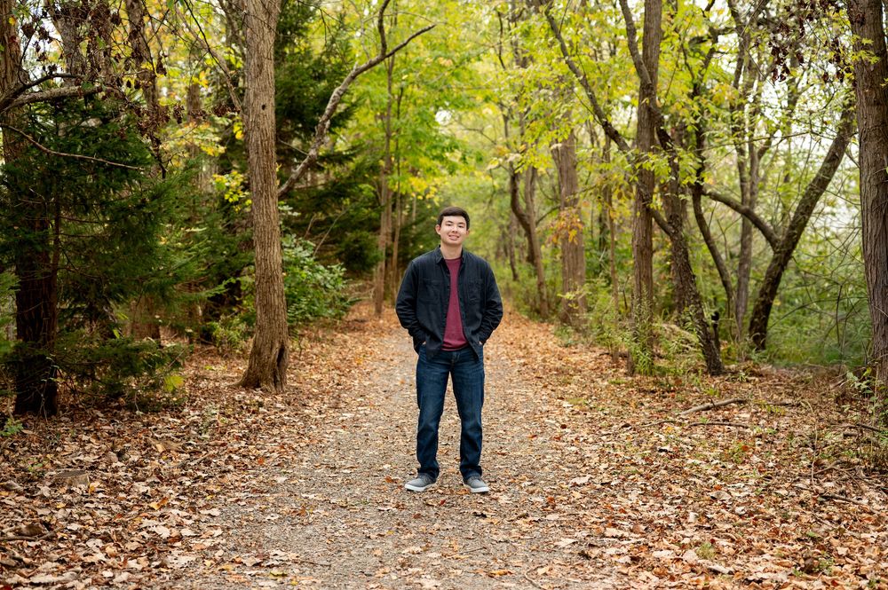 Young Asian man in dark jacket stands on leaf-covered forest path surrounded by autumn trees.