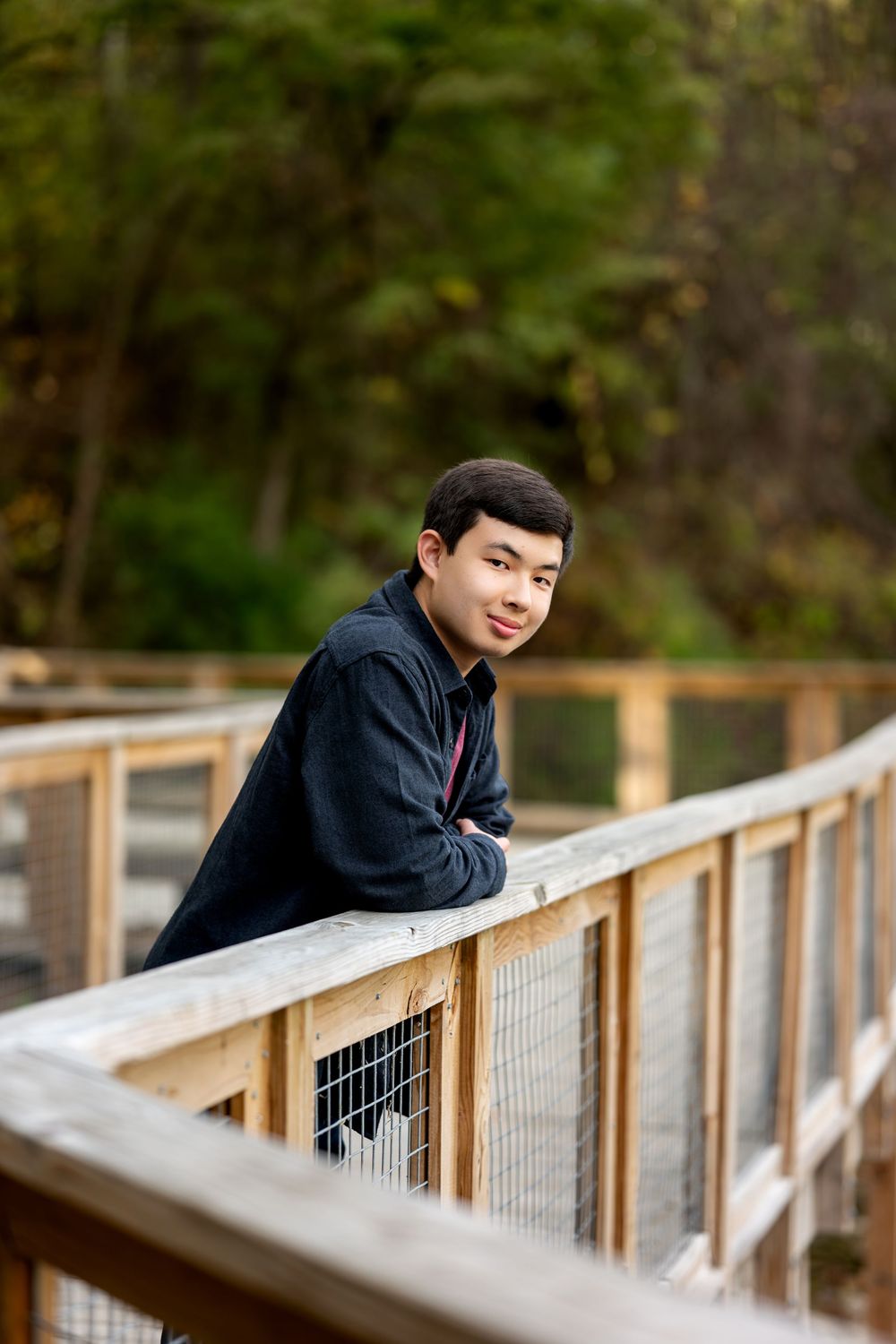 Young Asian teen smiling on wooden bridge railing in autumn park.