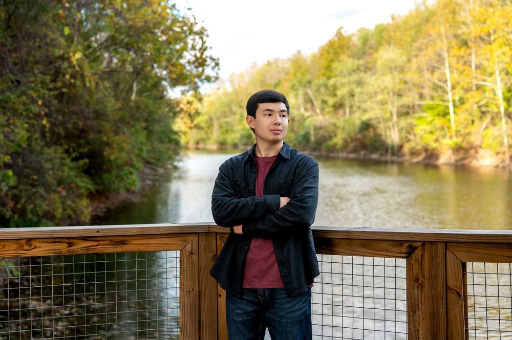Young Asian man leaning on railing gazing at peaceful river view.