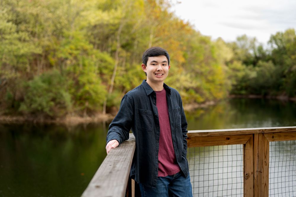 Smiling young Asian man in red shirt and dark jacket leans on wooden bridge railing by river.