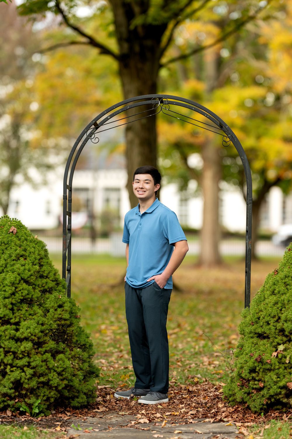 Smiling young man in blue polo stands under metal garden arch surrounded by autumn foliage in a park.