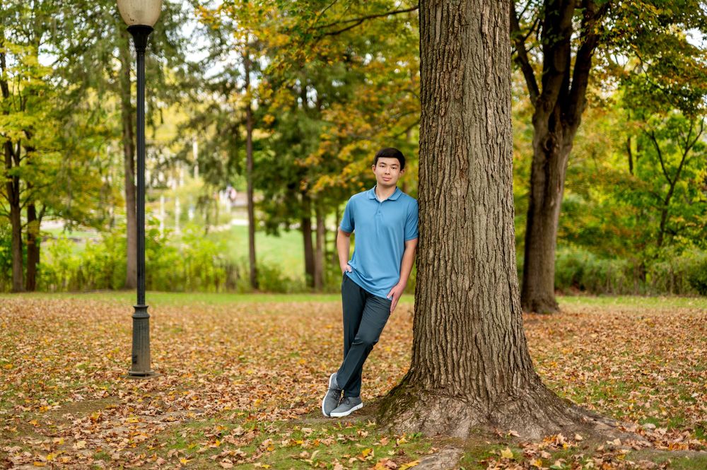 Young man in blue polo leans against large tree in a scenic autumn park with fallen leaves.