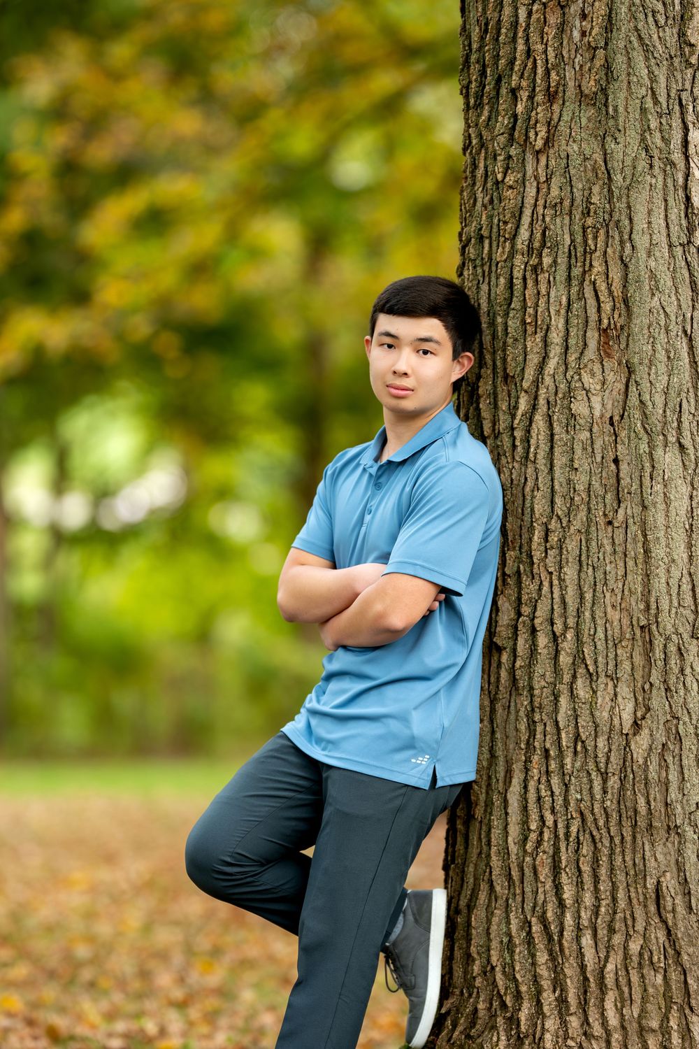 Teen boy in blue polo stands against tree with arms crossed and one foot raised in autumn park.
