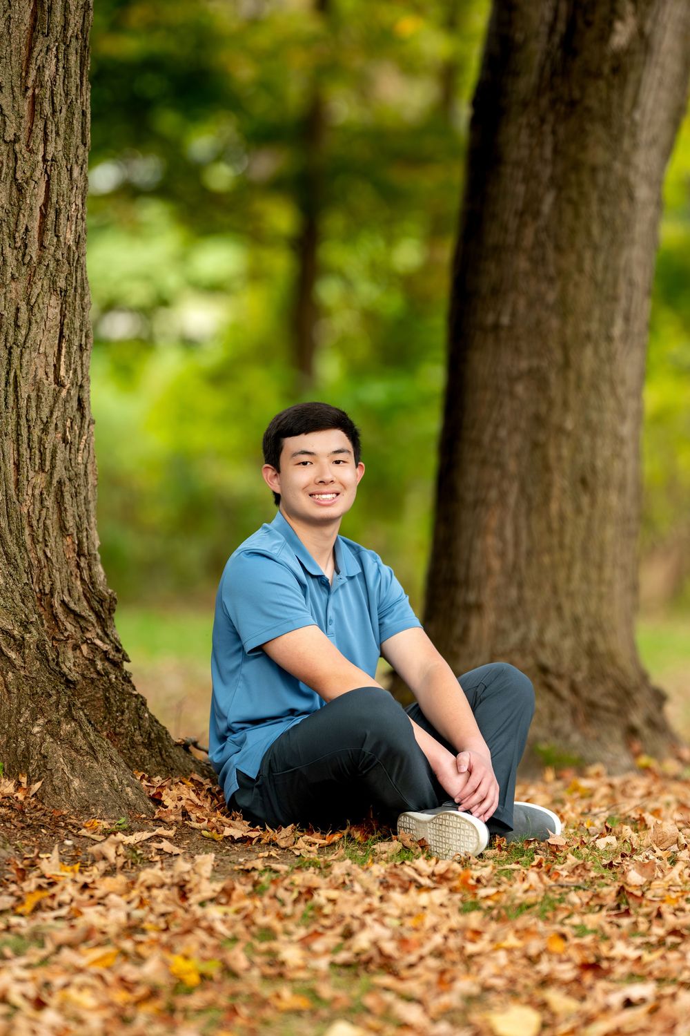 Smiling young man in blue polo sits cross-legged among fallen leaves between two large trees.