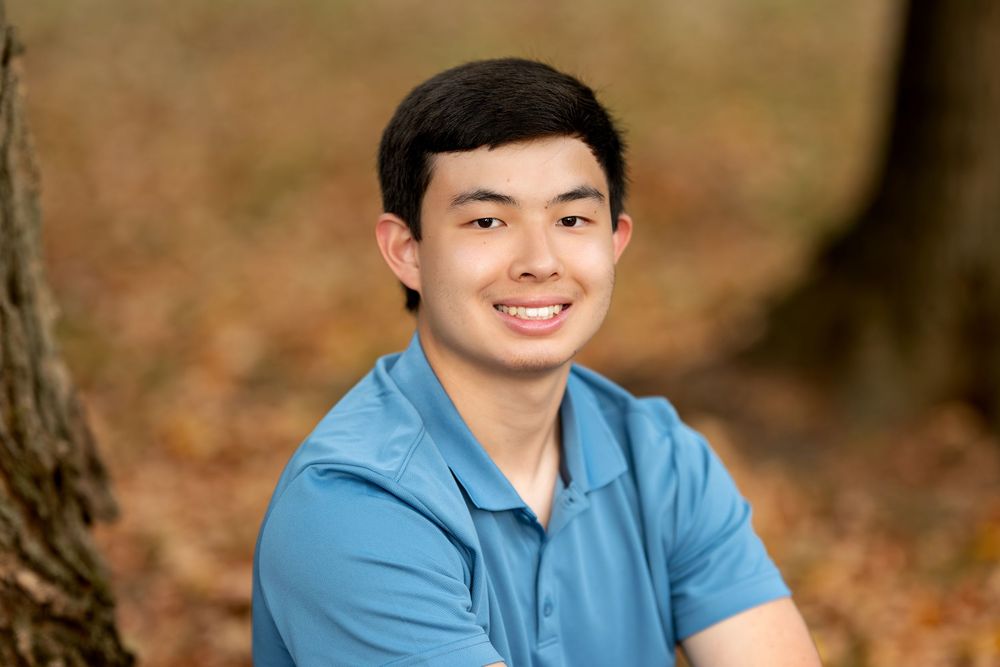 Close-up portrait of smiling young man in blue polo shirt outdoors with autumn leaves background.