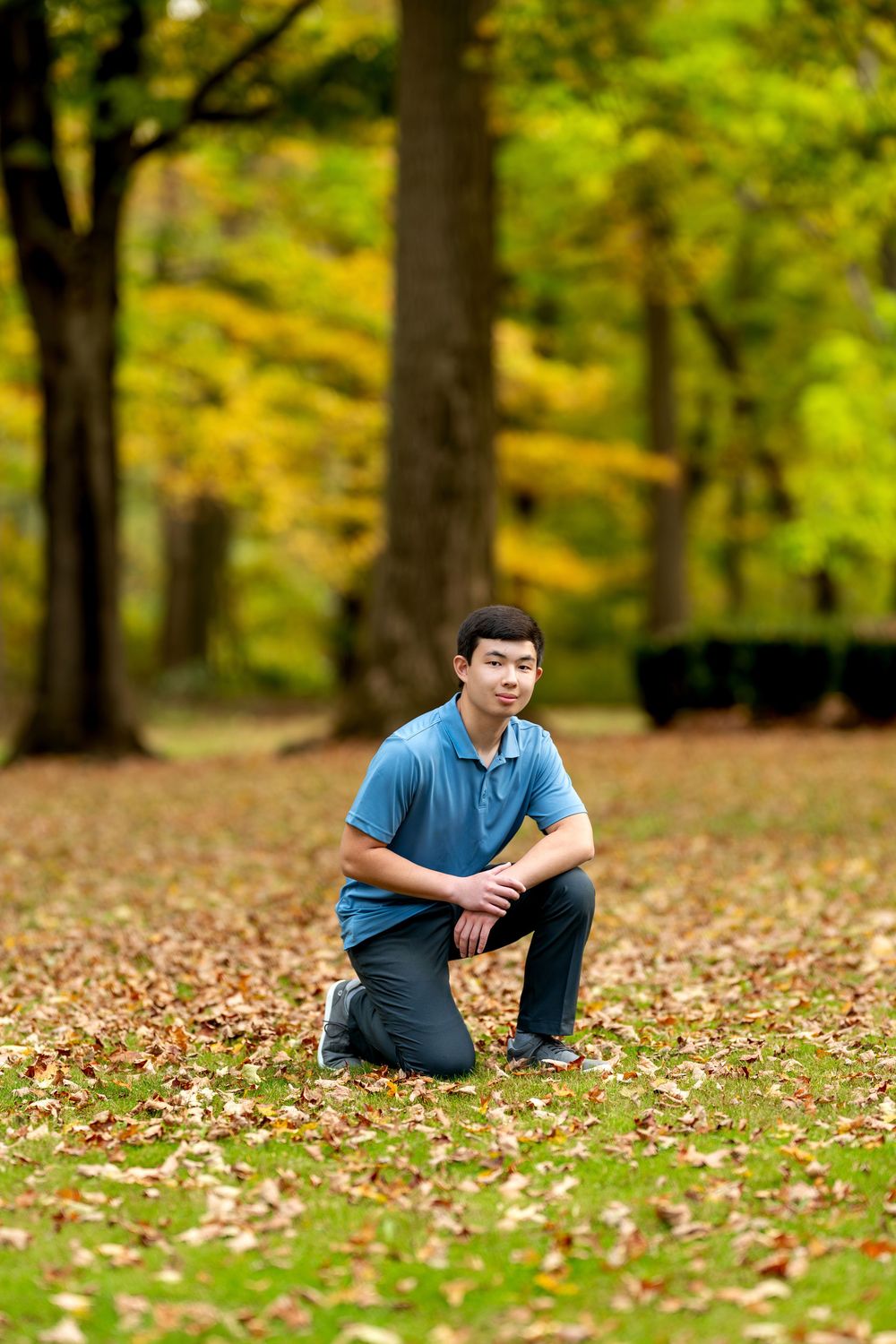 Young Asian man in blue polo kneeling on ground covered in fallen autumn leaves in a park.