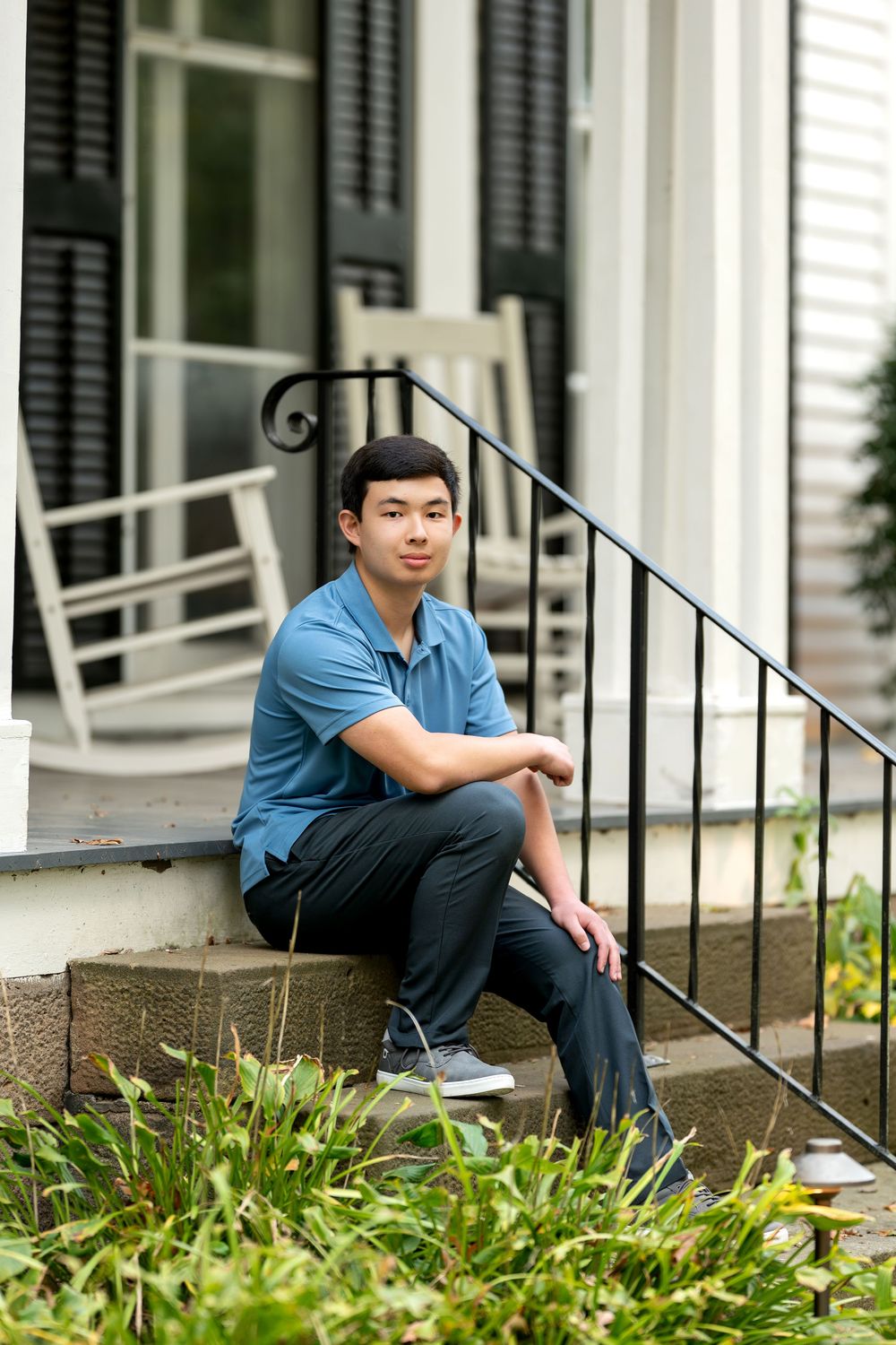 Young Asian man in blue polo shirt sitting on front porch steps of white house with rocking chairs.