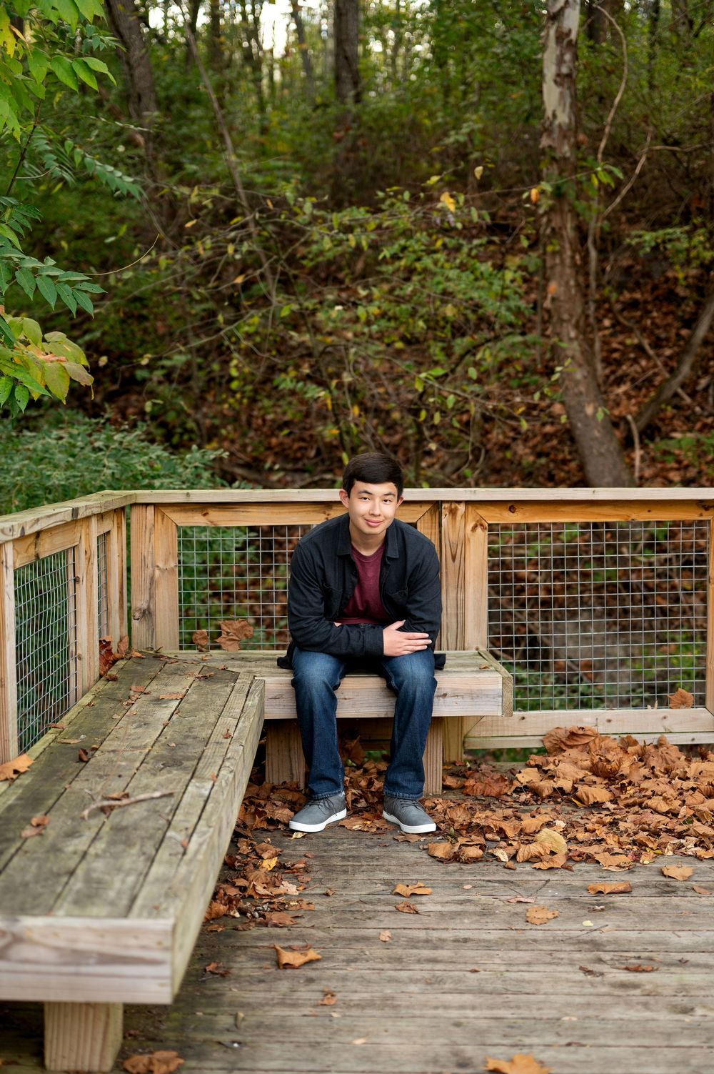 Young Asian teen sitting on wooden boardwalk bench surrounded by colorful fall foliage in forest.