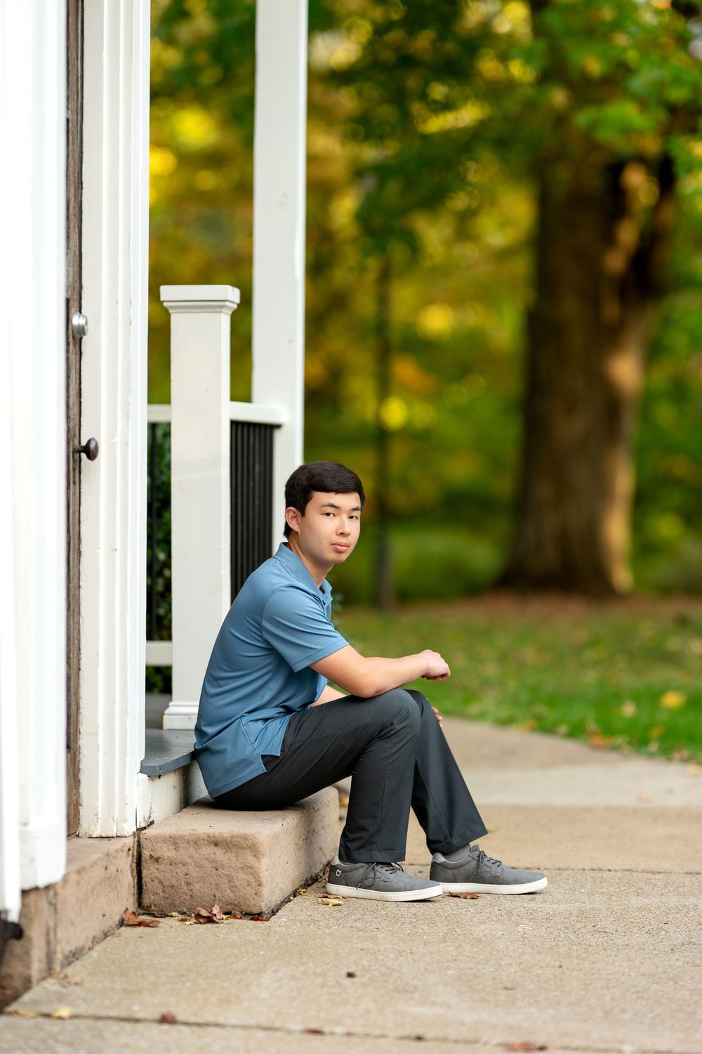 Young Asian man in blue polo shirt sitting on porch steps with autumn trees in background.