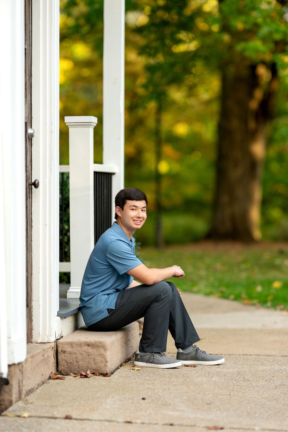 Smiling young Asian man in blue polo shirt sitting on porch steps with colorful autumn trees behind.