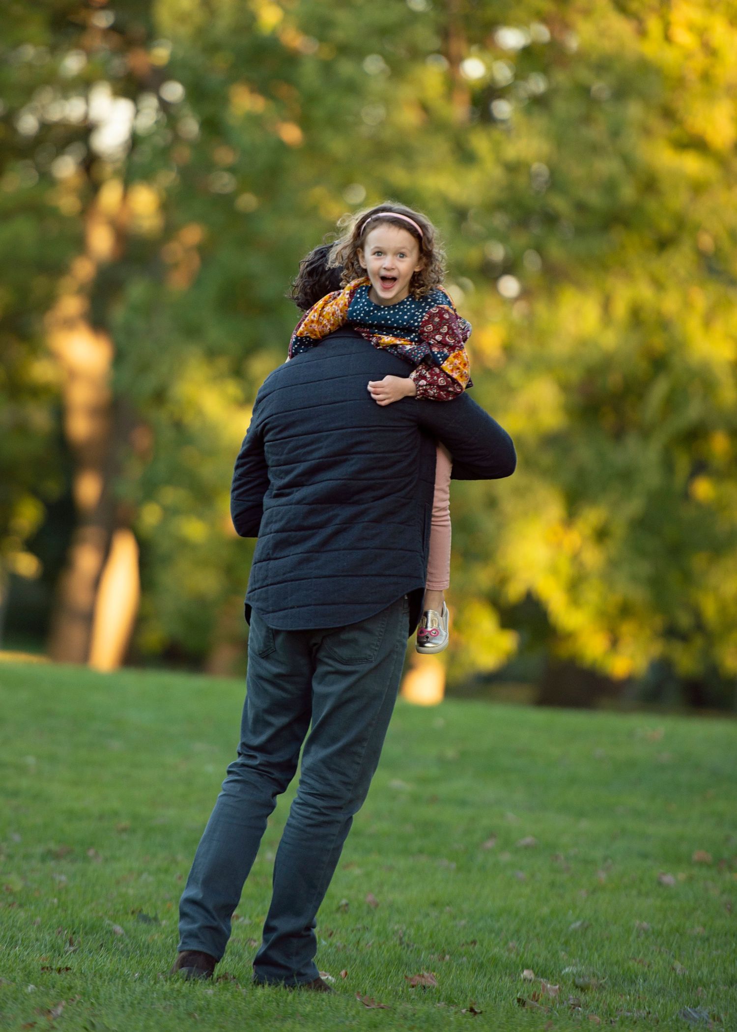 A man carries a laughing child on his back in a sunny autumn park with lush green and yellow trees.