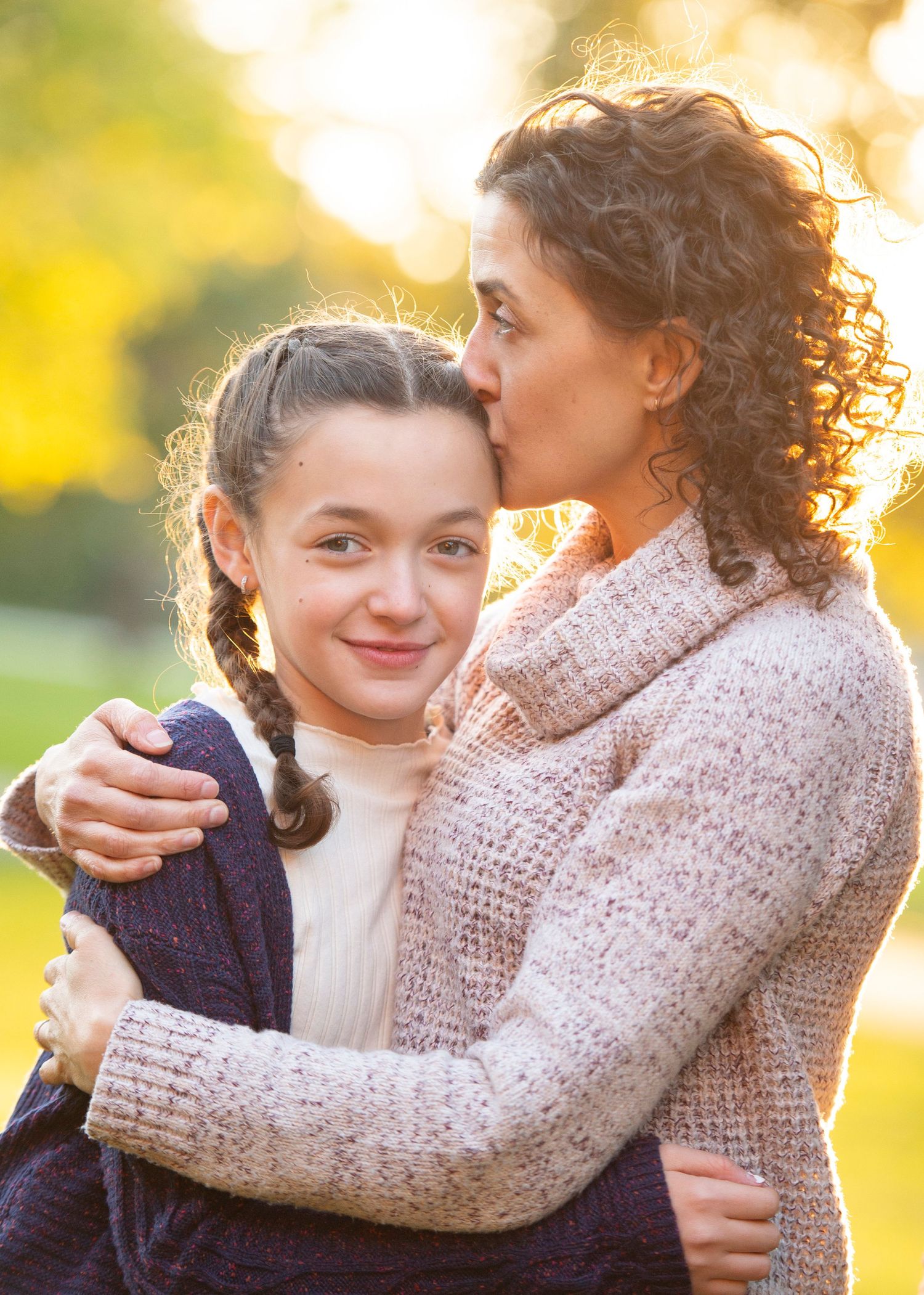 A mother with curly hair kisses her smiling daughter with braids outdoors in a sunny park setting.