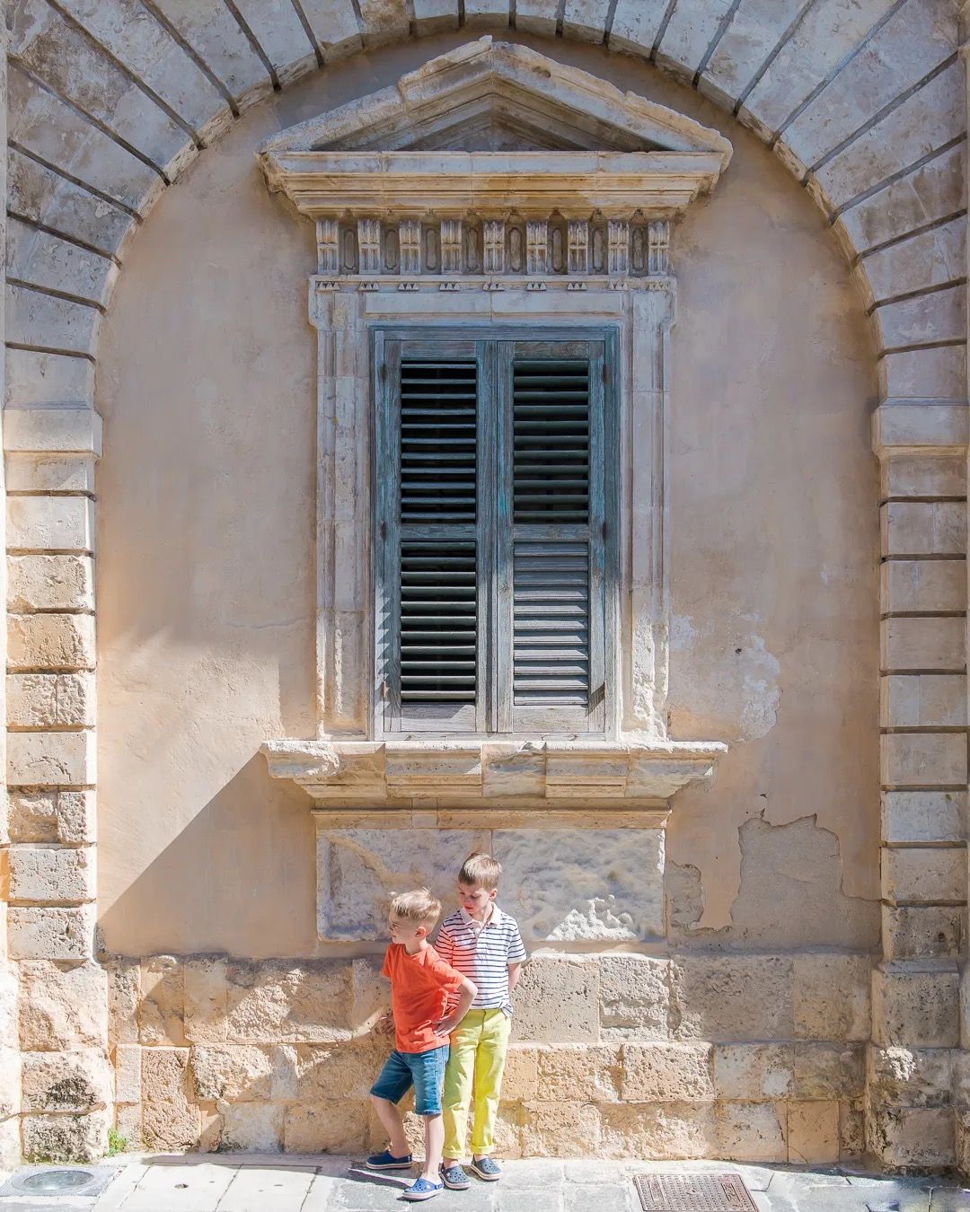 Young children stand beneath a tall stone building’s shuttered window in soft sunlight.