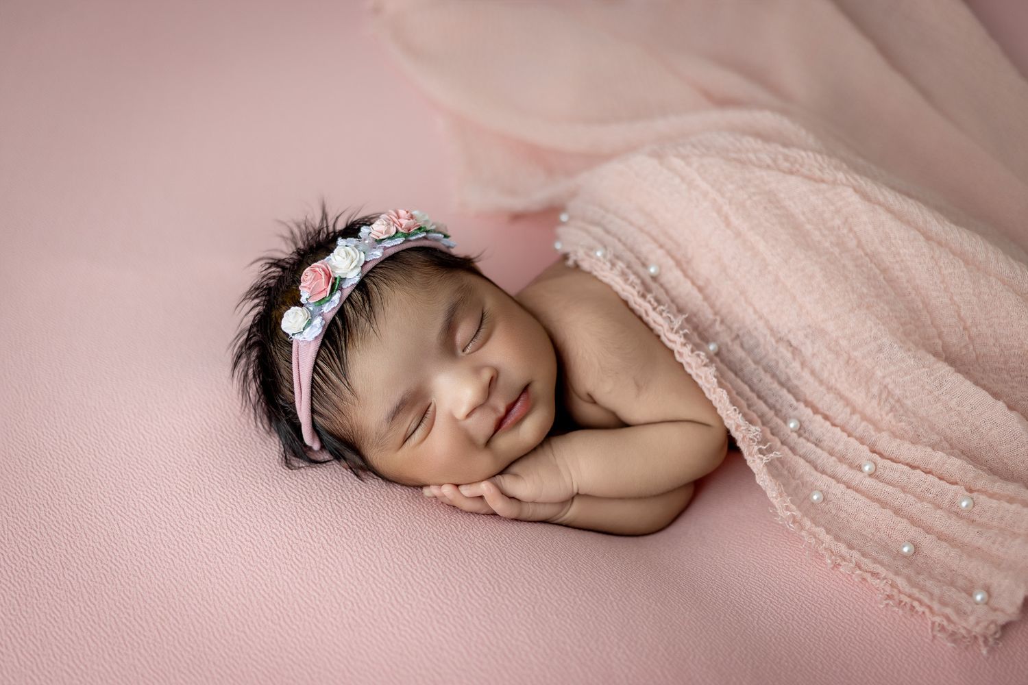 Sleeping newborn baby wearing a lace headband lies peacefully on soft pink fabric in a professional studio setting during newborn photoshoot