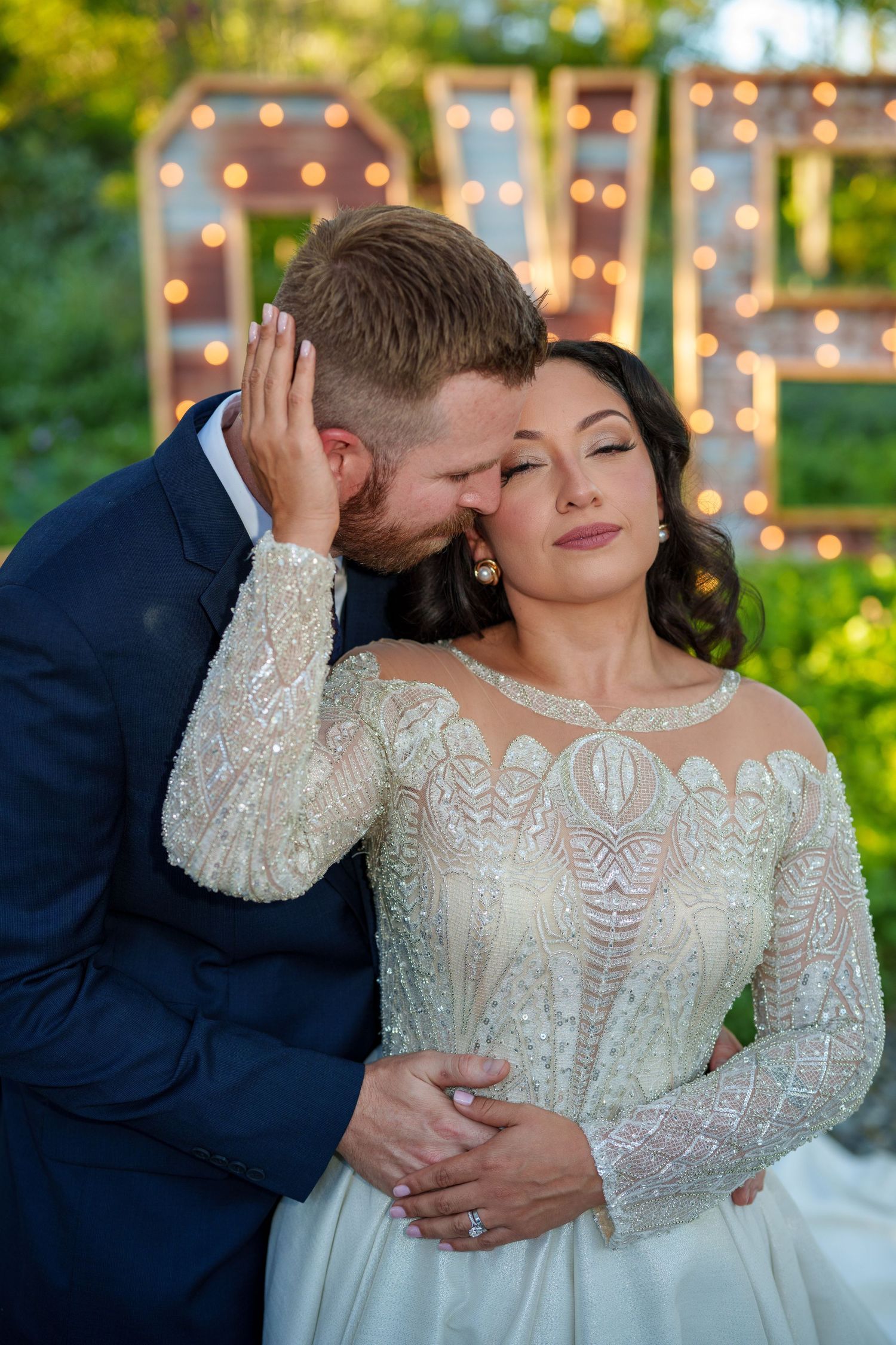 Romantic wedding couple embraces outdoors with twinkling lights in background creating intimate moments.
