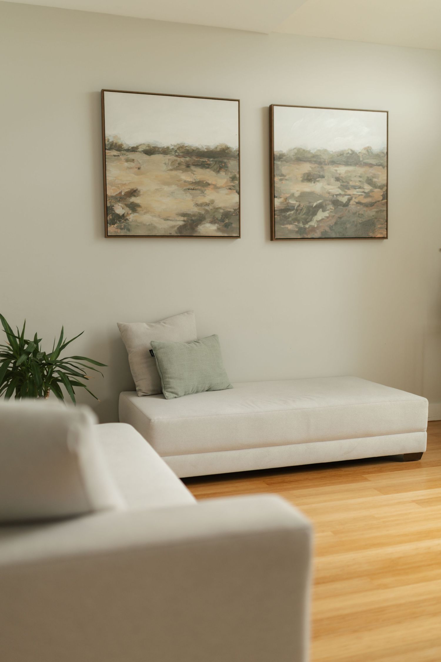 Minimalist living room with white daybed, sage green pillow, hardwood floors, and two landscape paintings.