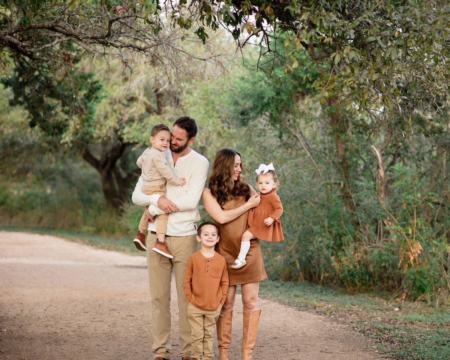 A family wearing coordinated earth-toned outfits walks together on a dirt path surrounded by green trees. Fall Mini Session Brushy Creek
