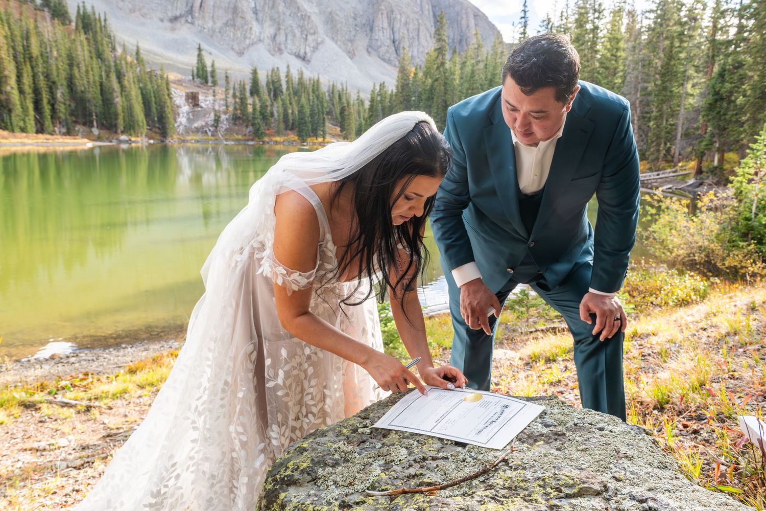 Bride and groom signing marriage certificate on a rock beside a mountain lake during an outdoor elopement ceremony.