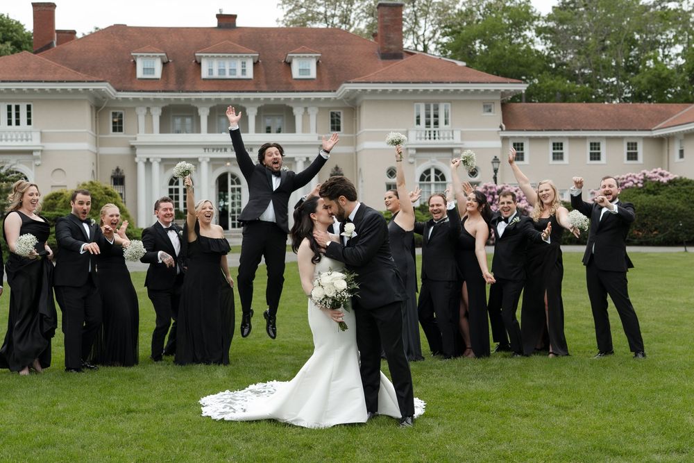Wedding couple kissing on lawn while bridal party in black celebrates behind them at elegant white mansion venue.