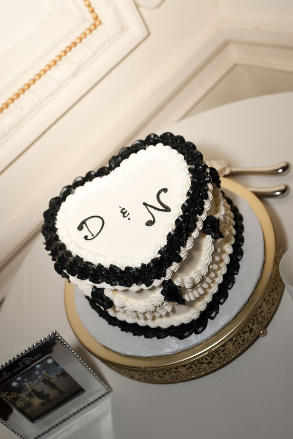Black and white heart-shaped two-tier wedding cake with monogram initials D and N on gold cake stand.