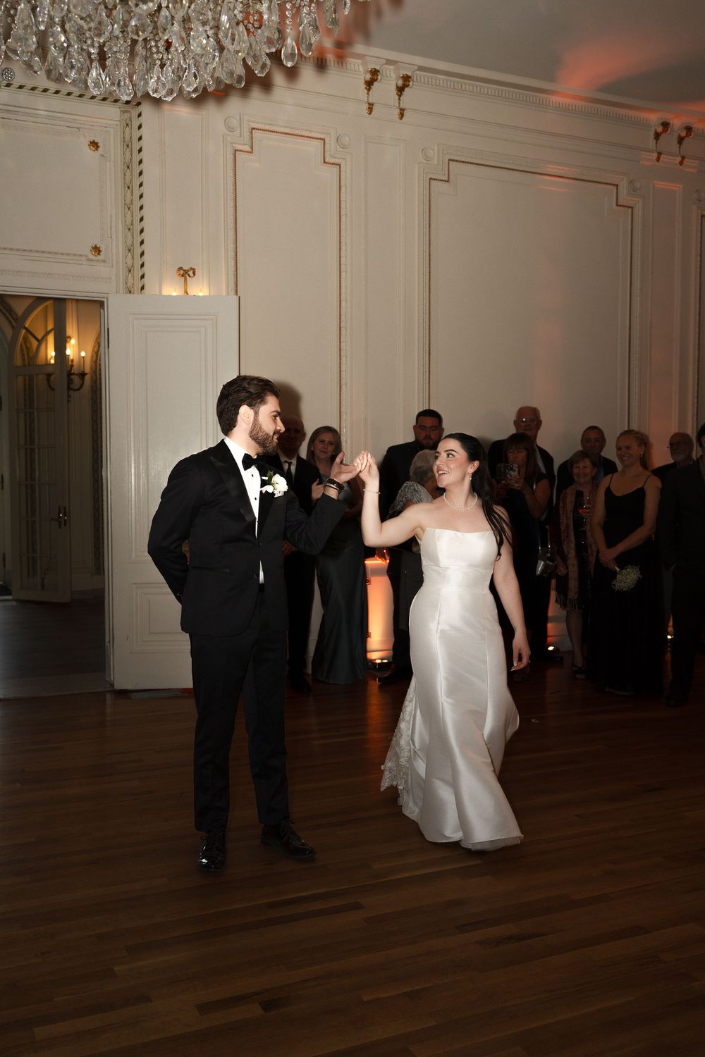 A bride and groom share their first dance in an elegant ballroom while wedding guests watch in the background.