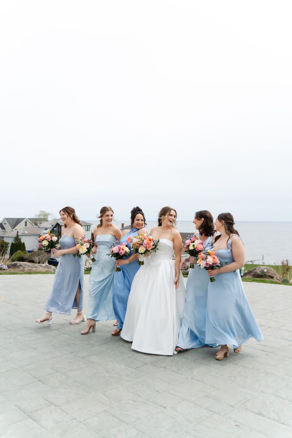 Bride in white gown walks with five bridesmaids in light blue dresses holding colorful bouquets near waterfront.