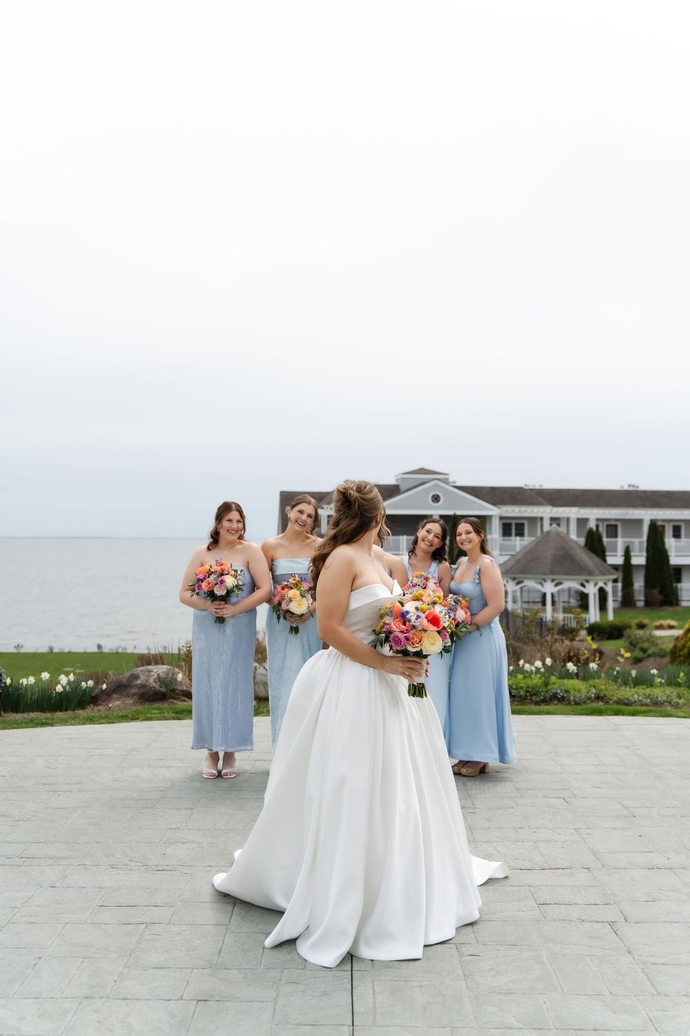 Bride in white gown holds colorful bouquet with bridesmaids in light blue dresses near waterfront venue.