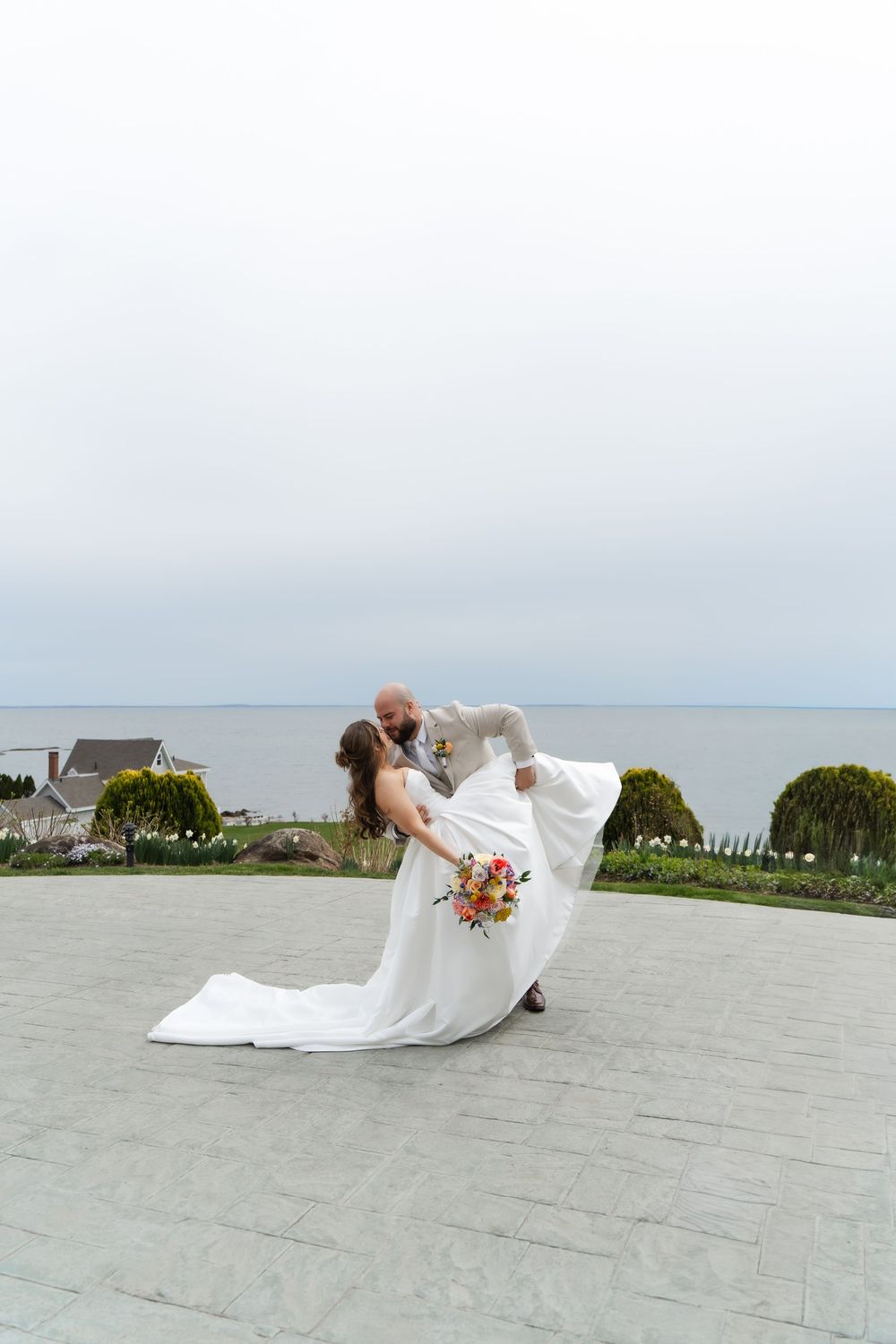 Groom dips bride in white gown with bouquet, oceanview backdrop on stone terrace.
