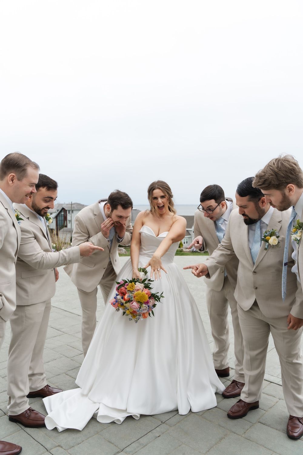 Bride in white gown holds colorful bouquet surrounded by groomsmen in tan suits on outdoor terrace.
