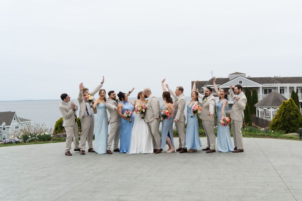 Wedding party celebrating outdoors by the ocean, bridesmaids in blue dresses, groomsmen in tan suits, bride in white gown.