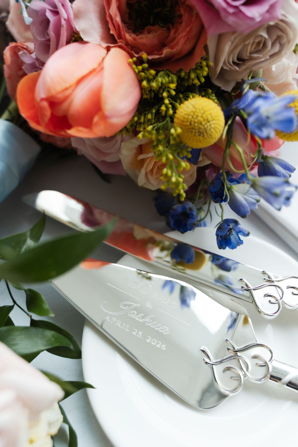 Engraved silver wedding cake server set beside colorful bridal bouquet with pink blooms.