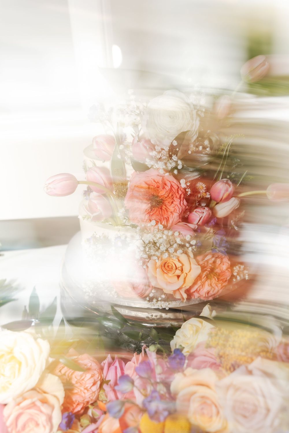 Elegant wedding cake adorned with pink tulips, coral roses, and baby's breath flowers in a dreamy, soft-focus style.