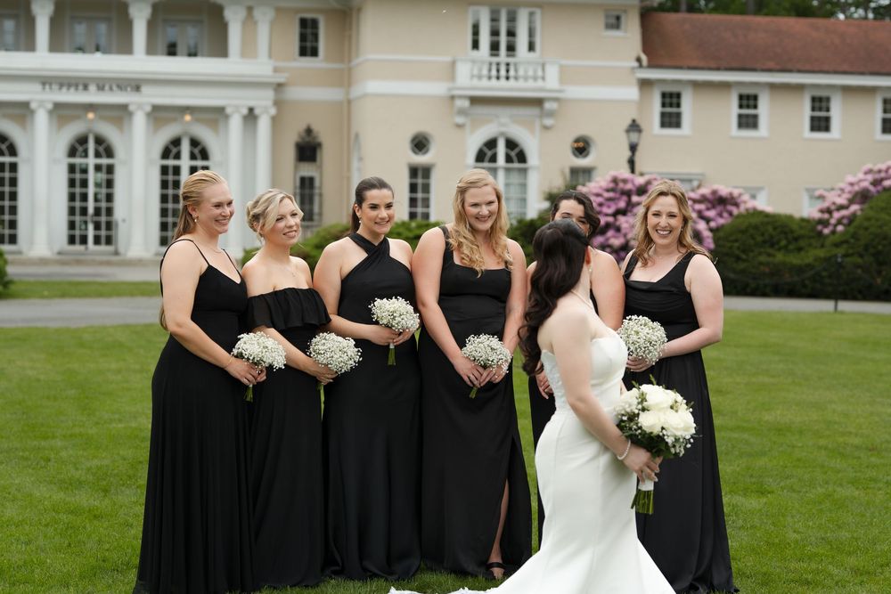 Bride in white gown with bridesmaids in black dresses holding white bouquets outside elegant mansion venue.