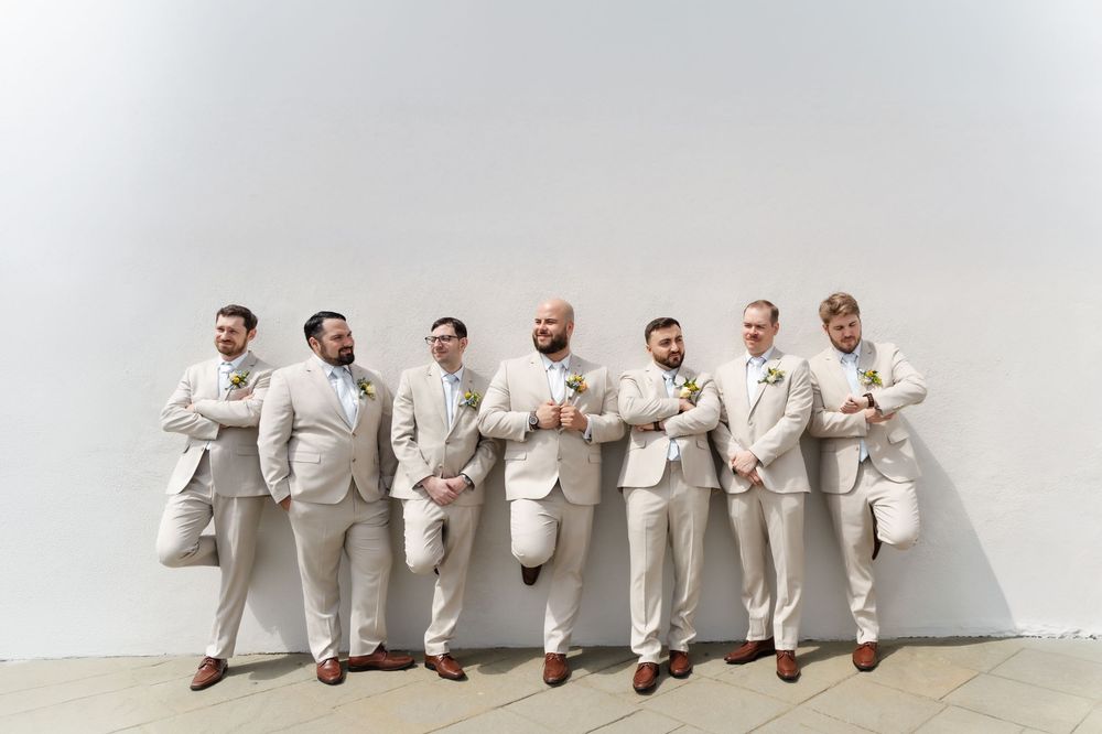 Seven groomsmen in matching tan suits with boutonnieres pose casually against a white wall at a wedding.