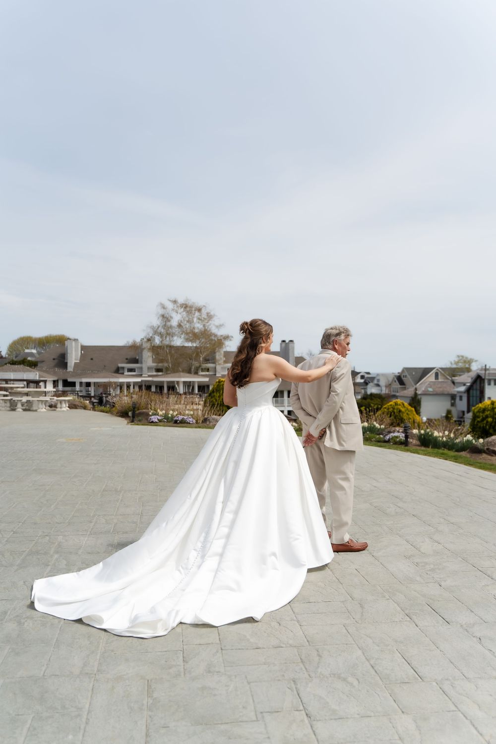 Bride in a flowing white ball gown stands with an older woman in a beige suit on a stone patio at a wedding.