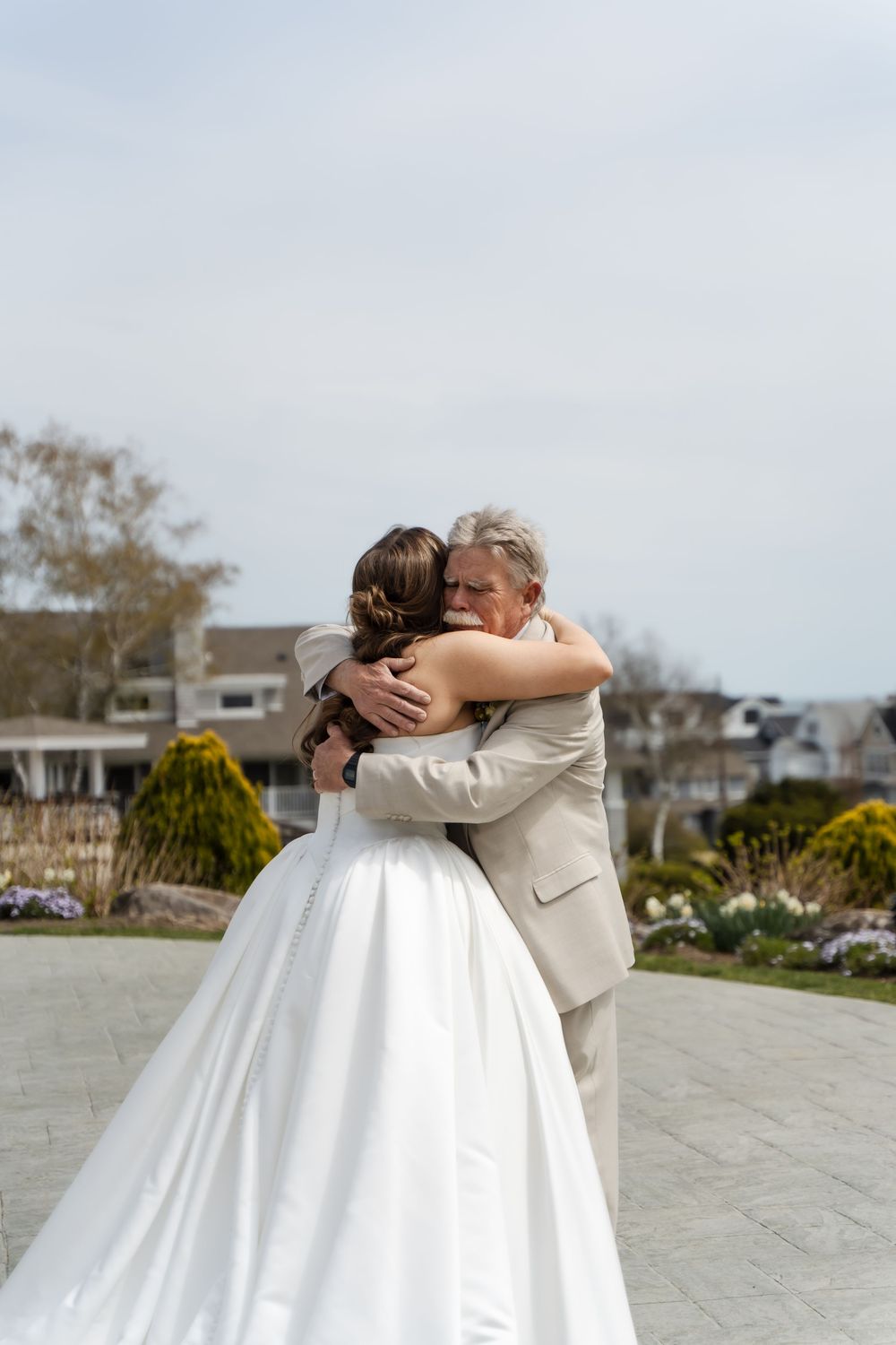 Bride in white gown shares emotional hug with older man in tan suit outdoors at wedding venue.