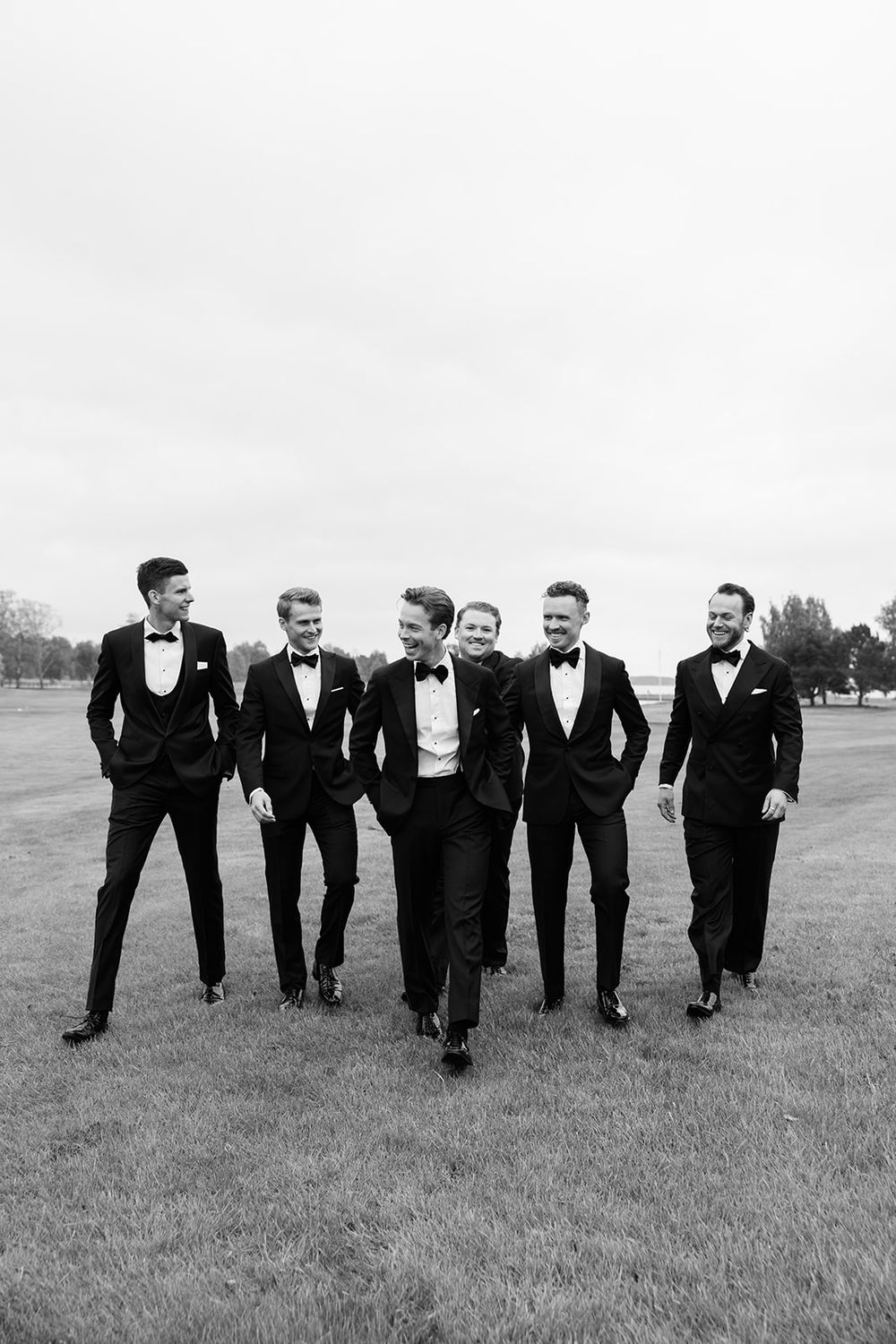 Six groomsmen in black tuxedos and bow ties walk and laugh together on a green lawn in a black and white photo.