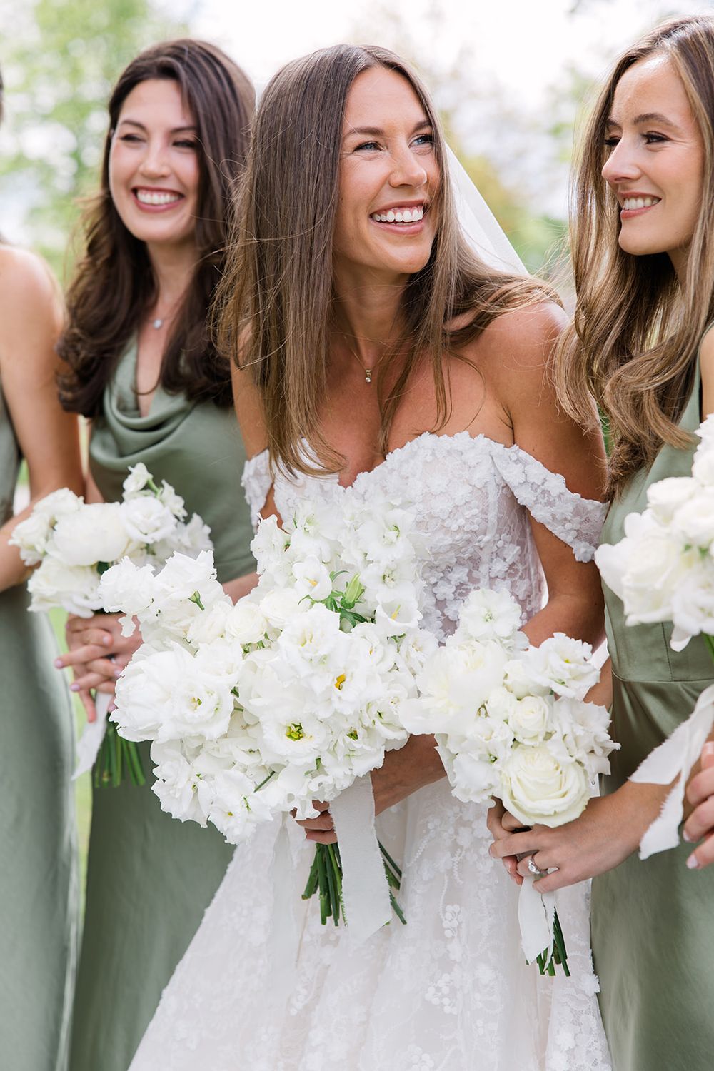 Bride in white lace gown holds bouquet with bridesmaids in sage green dresses, all smiling and holding white floral bouquets.