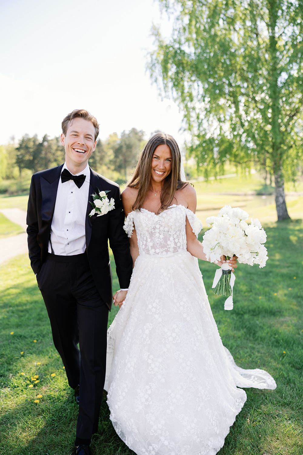 Happy bride and groom in formal wedding attire laughing together outdoors on green lawn with white bridal bouquet.