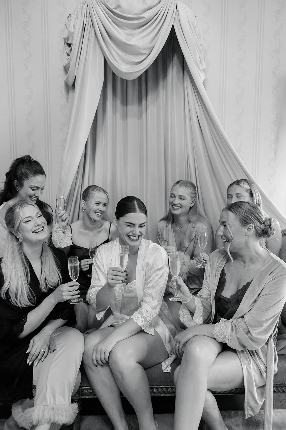 Black and white photo of a bride and bridesmaids in robes toasting with champagne glasses during a bridal party celebration.
