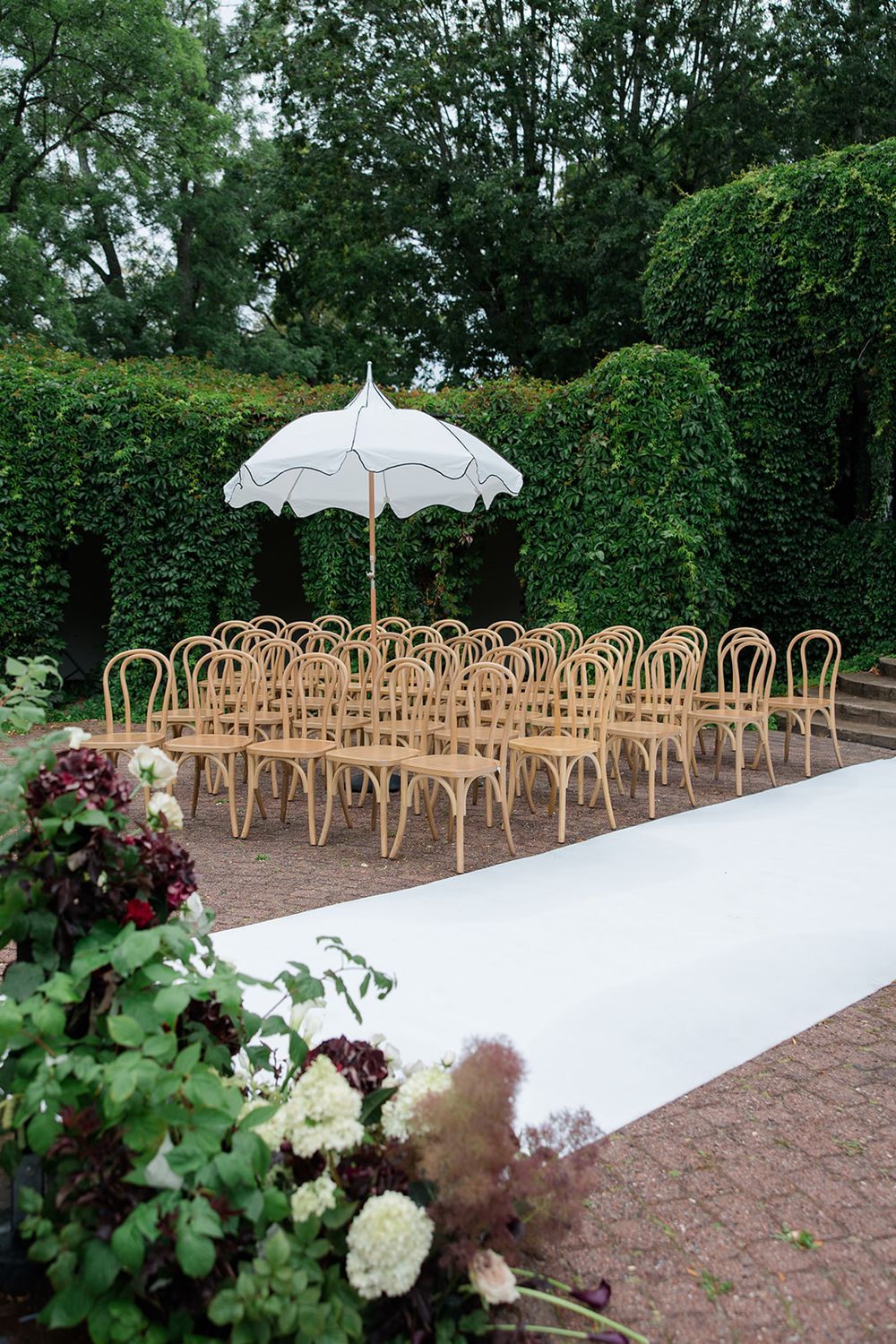 Elegant outdoor wedding ceremony setup with rattan chairs, white aisle runner, parasol, and lush green hedge backdrop.