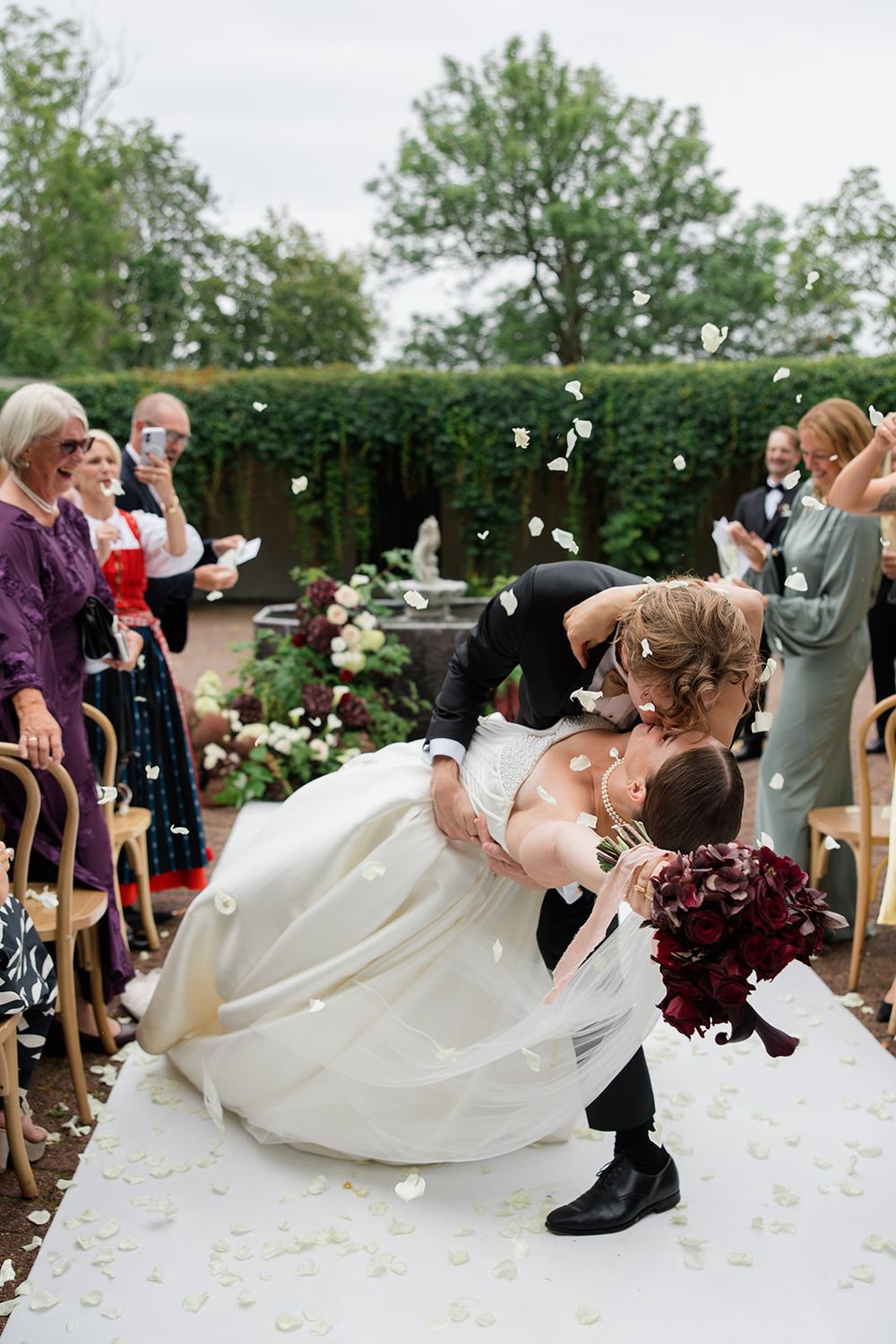 Groom dips bride for a kiss at outdoor wedding ceremony while guests toss white flower petals.