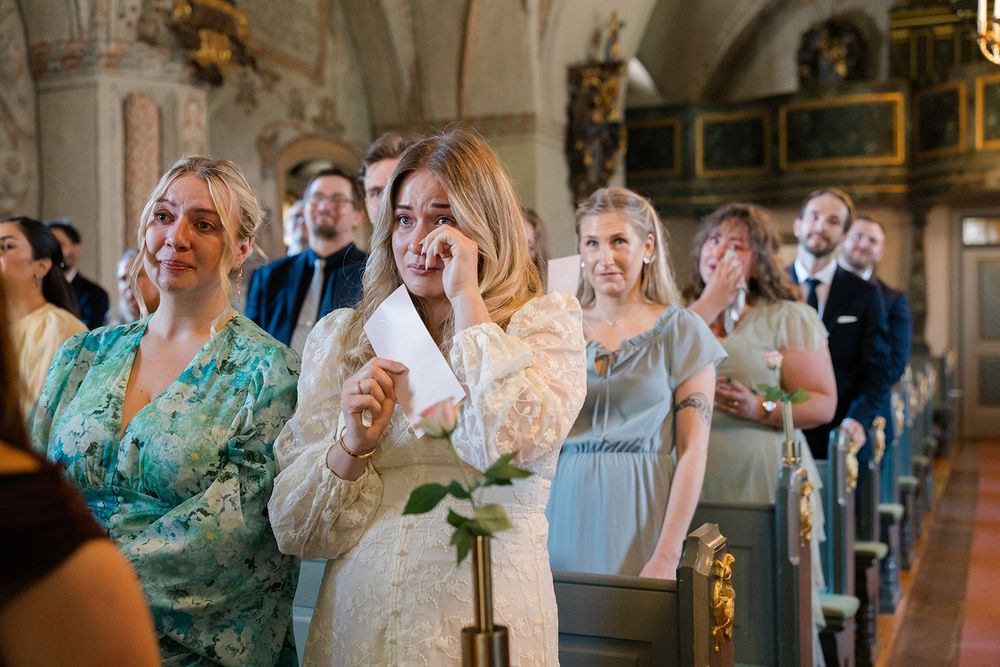 Emotional wedding guests wiping tears in a church aisle during a ceremony, dressed in elegant formal attire. Bröllop Hedenlunda Slott