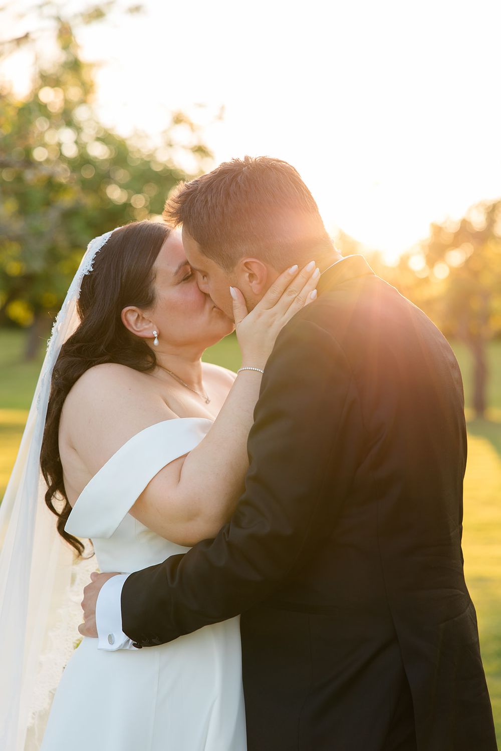 A bride and groom share a romantic kiss at golden hour, bathed in warm sunset light on a lush green lawn. Bröllop Hedenlunda Slott