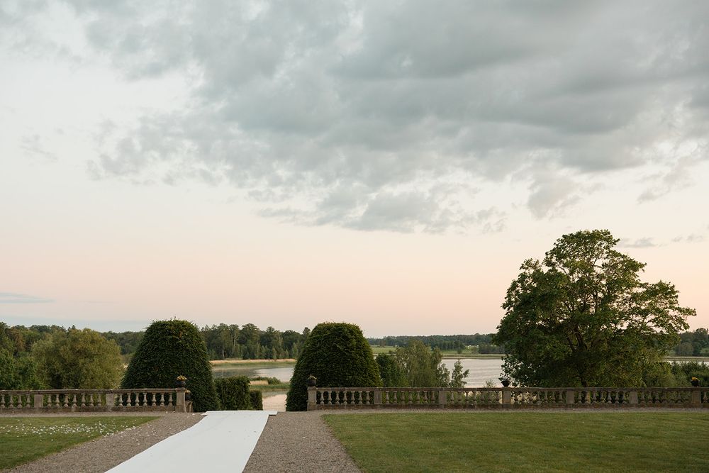 Elegant garden terrace with manicured hedges and a serene lake view under a dramatic cloudy sky. Bröllop Hedenlunda Slott