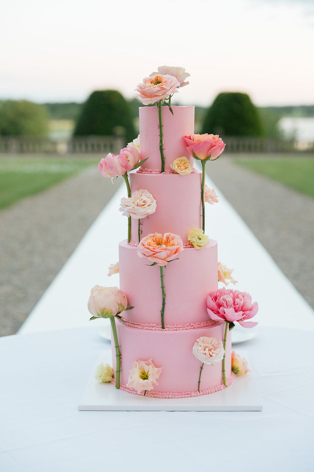 Elegant four-tier pink wedding cake adorned with fresh pink and peach flowers on an outdoor table. Bröllop Hedenlunda Slott Bröllopstårta