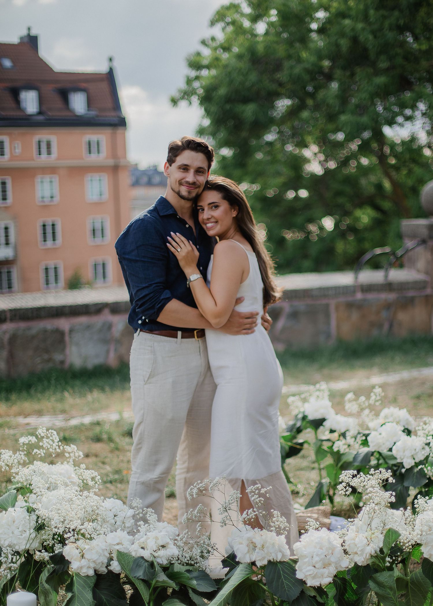 Happy couple embracing and smiling surrounded by white floral arrangement during outdoor marriage proposal setup.